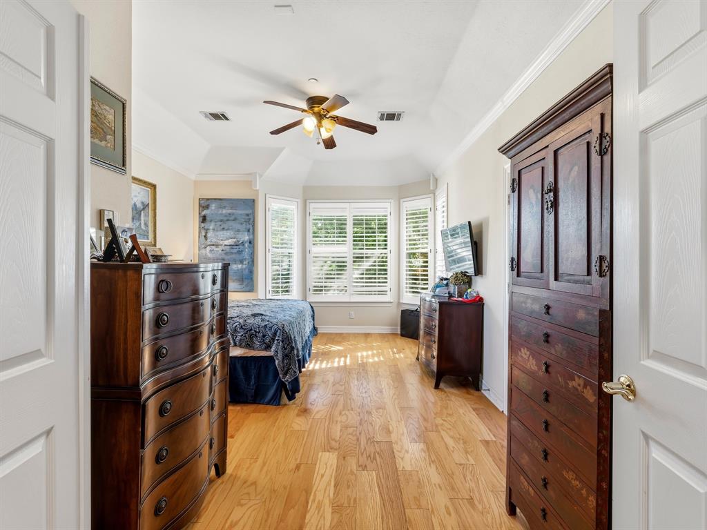 Tray ceiling with a central multi-blade ceiling fan and wood-finish flooring, complemented by a bay window featuring white plantation shutters