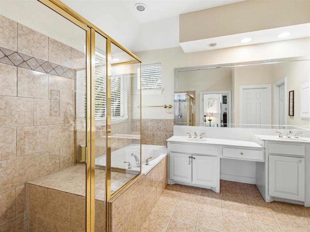 Bathroom featuring a dual-sink vanity with white cabinetry, a glass-enclosed shower with tiled walls and gold-tone trim, and a deep soaking tub