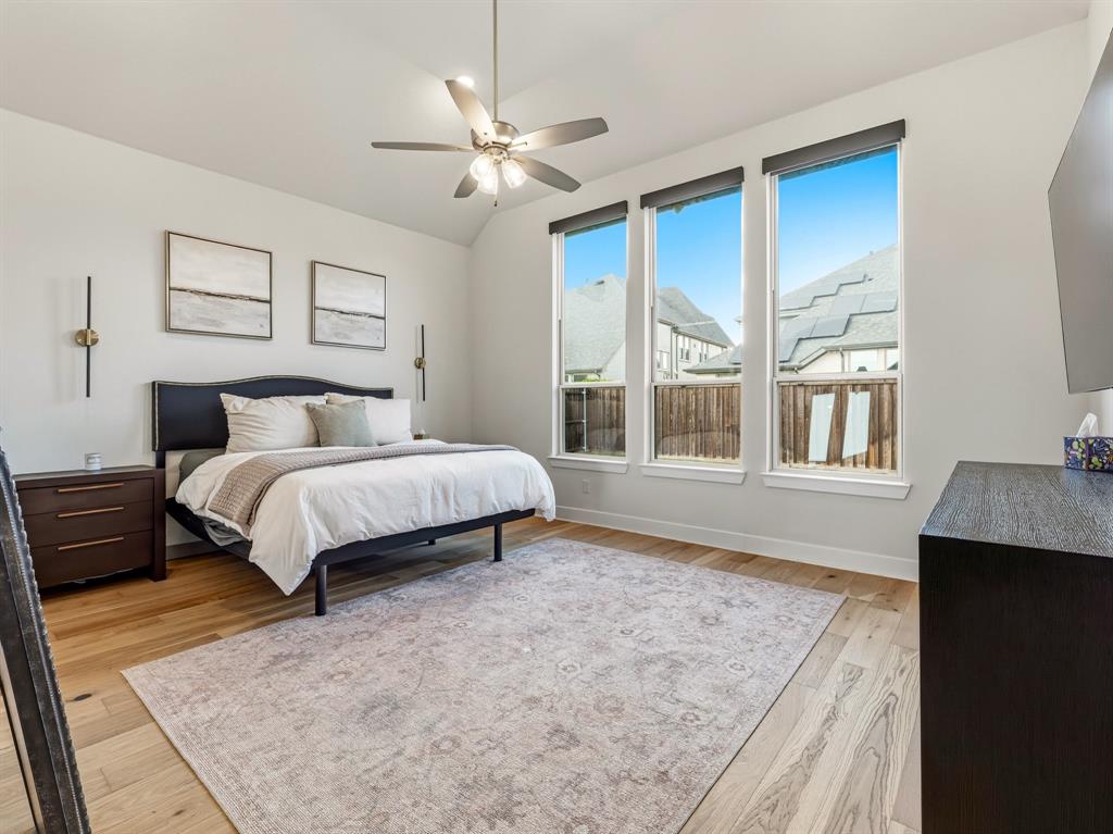 Bedroom featuring vaulted ceilings, wood-finish flooring, a central ceiling fan with integrated lighting, and three tall windows with sleek roller shades