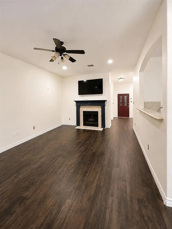 Living area featuring wood-finish flooring, a fireplace with a mantel and tile surround, a multi-blade ceiling fan, and a pass-through opening with a ledge Living area featuring wood-finish flooring, a fireplace with a mantel and tile surround, a multi-blade ceiling fan, and a pass-through opening with a ledge