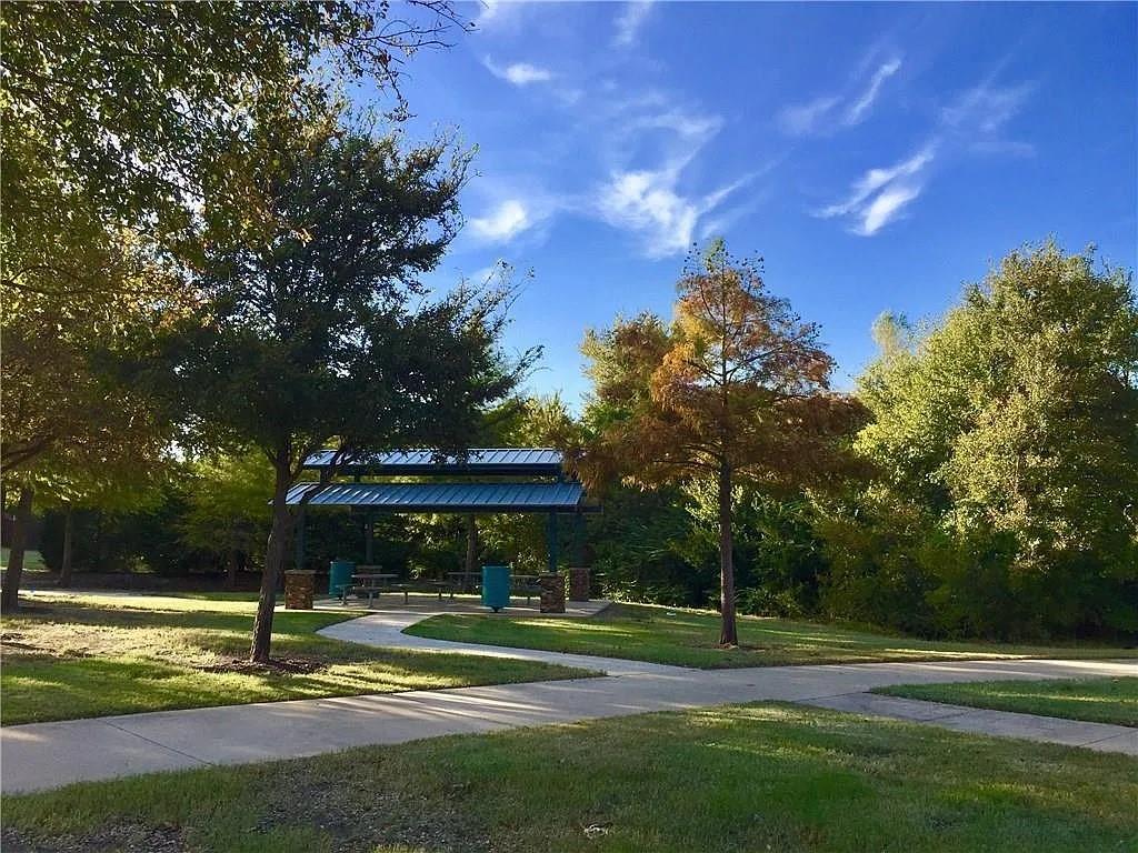 Outdoor pavilion featuring a metal roof and stone-clad pillars, situated on a concrete walkway amidst mature trees and landscaped grounds
