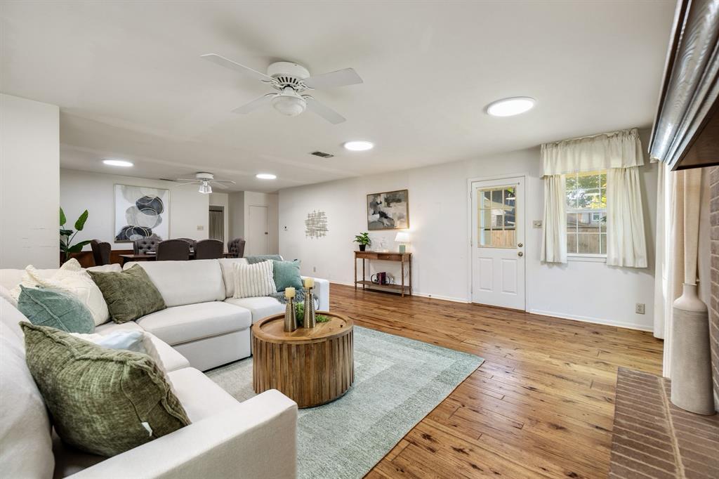 Open-concept living space featuring wood-finish flooring, recessed lighting, white-painted brick fireplace surround, dual ceiling fans, and an exterior door with glass pane detail