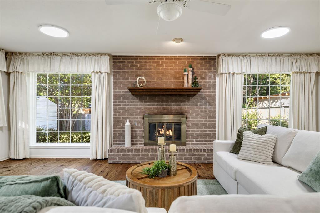 Floor-to-ceiling brick fireplace surround with a wooden mantel and metal firebox, wood-finish flooring, and dual picture windows with grid muntins