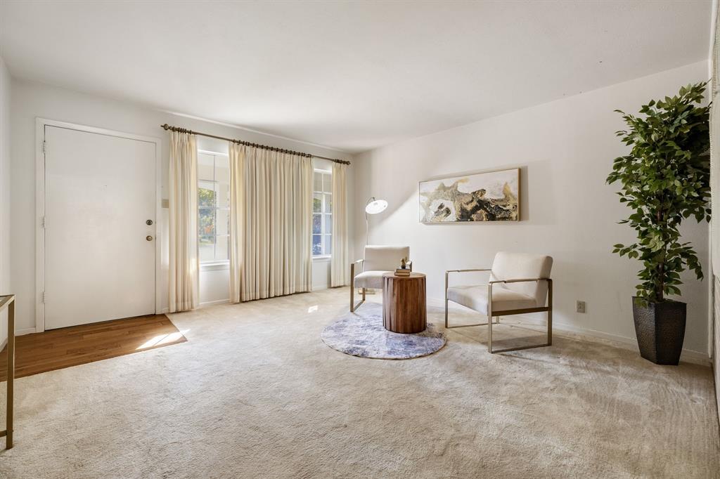 Neutral-toned interior space featuring light carpeting, wood-finish flooring at the entryway, and large windows with integrated window treatments Neutral-toned interior space featuring light carpeting, wood-finish flooring at the entryway, and large windows with integrated window treatments