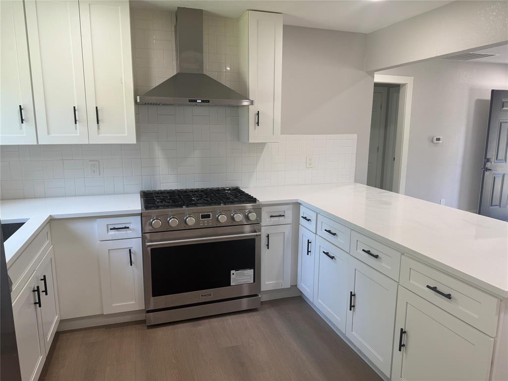 Kitchen featuring white shaker-style cabinetry with matte black hardware, stone-finish countertops, herringbone-pattern tile backsplash, stainless steel gas range, and stainless steel range hood