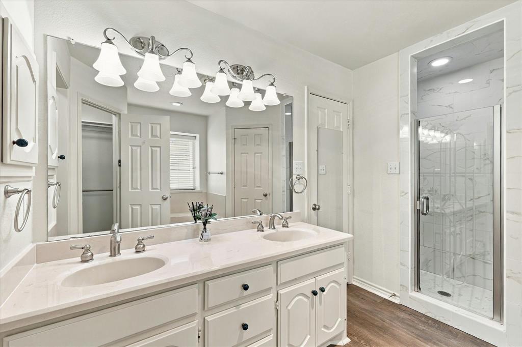 Dual-sink vanity with a light-toned countertop, large-format mirror, overhead multi-light fixture, and walk-in shower with stone-finish tile surround and glass door