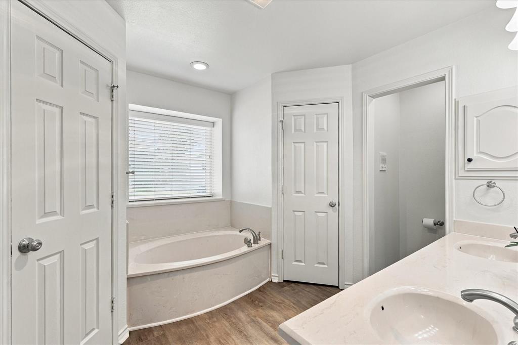Bathroom featuring a dual-sink vanity with a light-toned countertop, soaking tub with a deck surround, wood-finish flooring, and six-panel interior doors