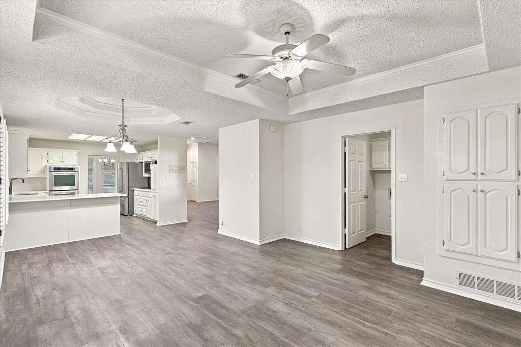 Interior area featuring wood-finish flooring, tray ceilings with crown molding, a central ceiling fan with integrated lighting, a built-in storage cabinet, and a kitchen equipped with a breakfast bar and stainless steel wall oven