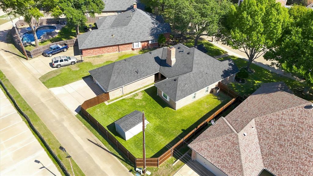 Aerial view of a corner lot property featuring a brick exterior, a shingle roof with a masonry chimney, a fenced-in backyard, and a paved driveway