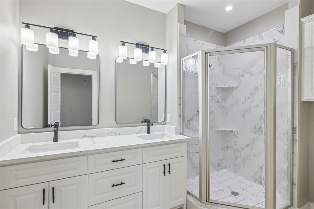 Dual-sink vanity with a solid-surface countertop and matte black fixtures, paired with two framed mirrors, multi-bulb wall lighting, and a walk-in shower featuring patterned tile surround and glass enclosure