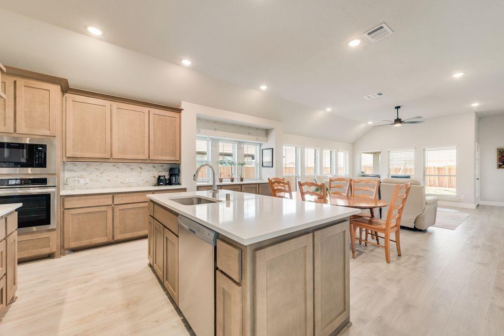 Kitchen featuring light wood-finish cabinetry, a central island with a solid-surface countertop, stainless steel appliances, and wood-finish flooring under a vaulted ceiling with recessed lighting