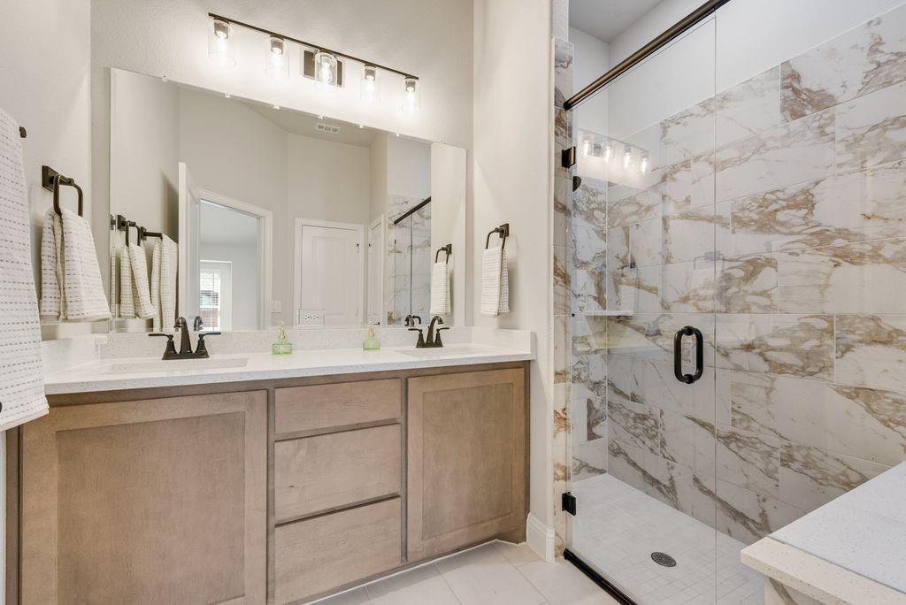 Double vanity featuring light-toned cabinetry, quartz countertops, dual under-mount sinks, matte black fixtures, and a walk-in shower with floor-to-ceiling stone-look wall tile and a glass enclosure