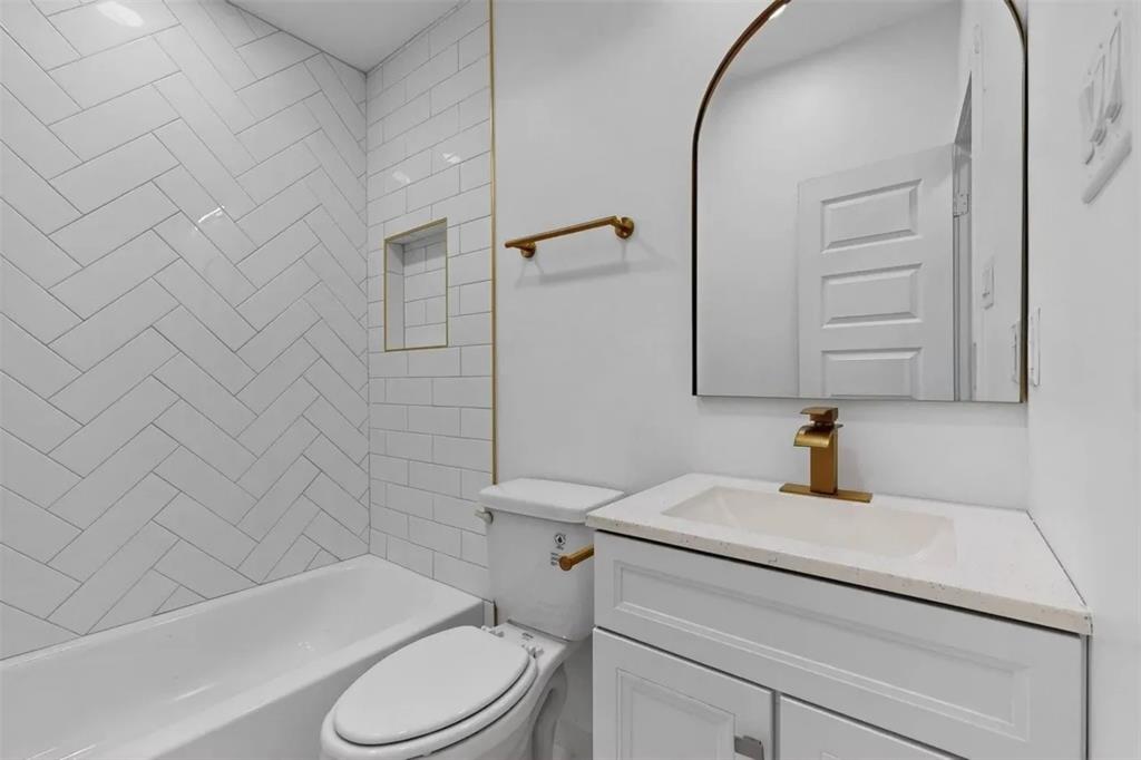 Bathroom featuring a herringbone-patterned tile tub surround, integrated wall niche, arch-top mirror, vanity with stone-composite countertop, and brushed-gold fixtures