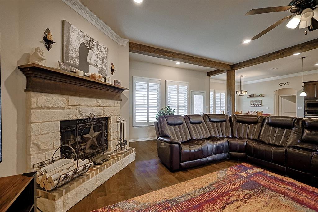 Stone-faced fireplace with a wood mantel, wood-finish flooring, ceiling fan with light kit, crown molding, and plantation shutters