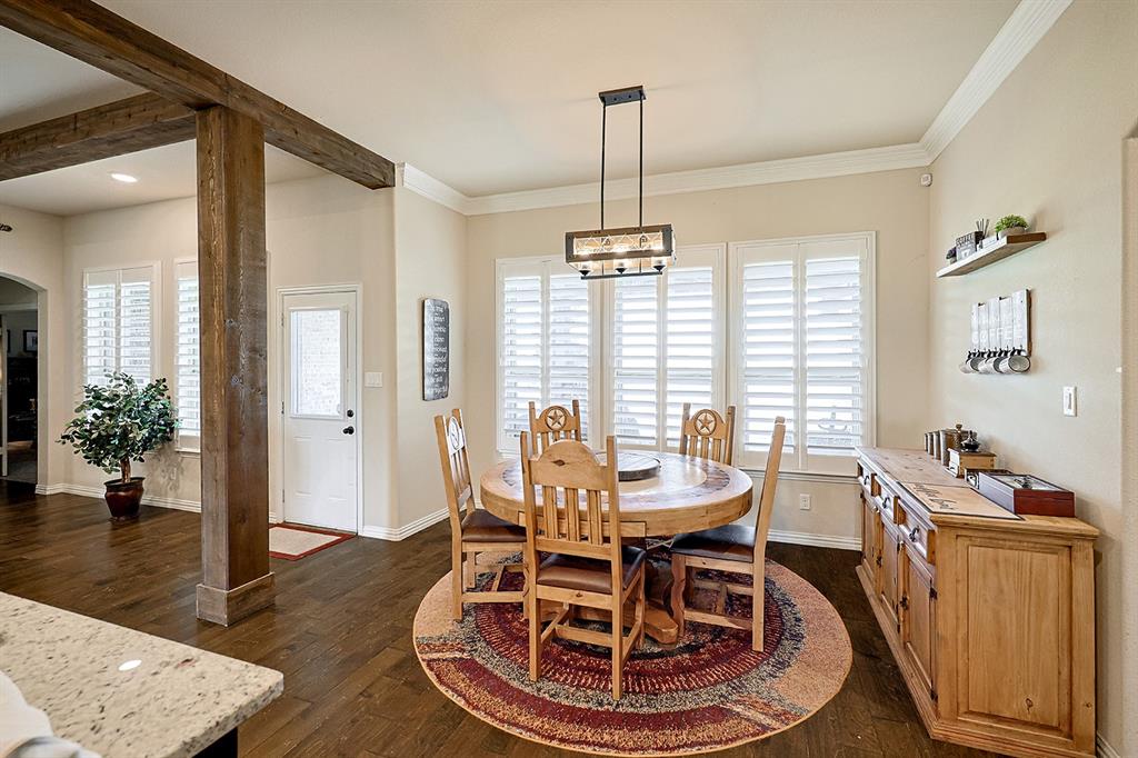 Dining space featuring wood-finish flooring, exposed stained-wood support beams, custom plantation shutters, crown molding, and a linear multi-light pendant fixture