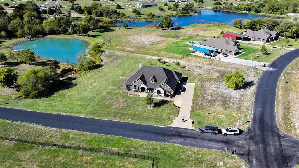 Aerial perspective of a multi-gabled residence featuring a paved driveway, exterior stone detailing, a pitched shingled roof, and an expansive lot with water features