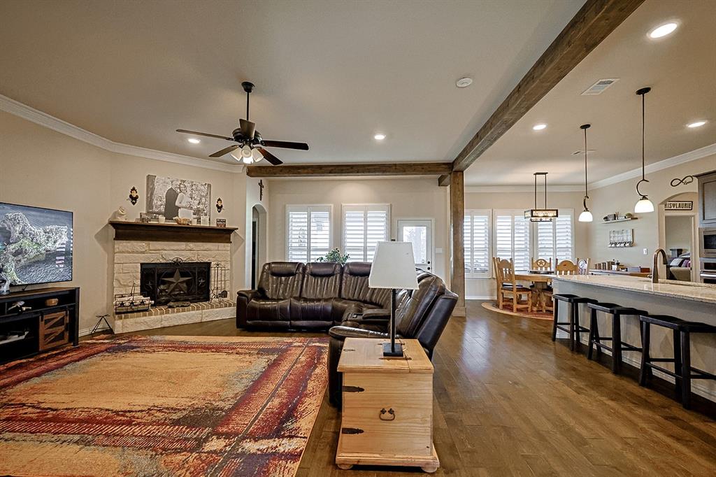 Open-concept interior featuring wood-finish flooring, exposed ceiling beams, a floor-to-ceiling stone fireplace, plantation shutters, and a kitchen island with pendant lighting
