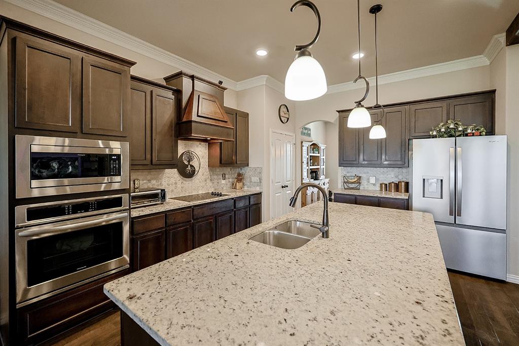 Kitchen featuring wood cabinetry, stone countertops, a central island with a double-basin stainless steel sink, built-in stainless steel double wall ovens, and pendant lighting