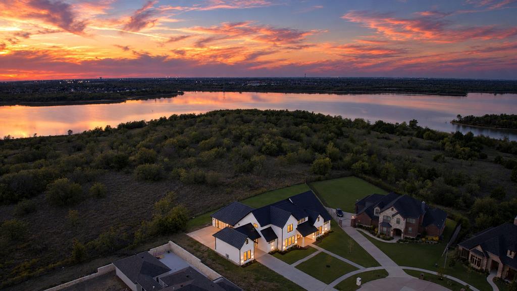 Aerial perspective of a multi-gabled residence featuring a light-toned exterior, dark-toned roof, concrete driveway, and direct proximity to a waterfront and natural landscape