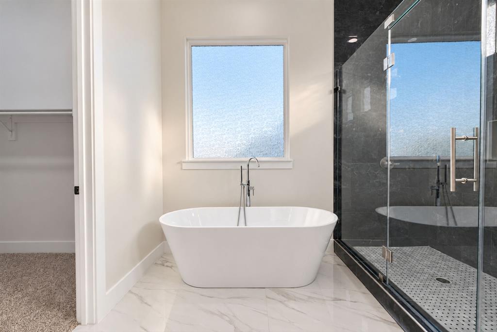 Freestanding soaking tub with a floor-mounted faucet, walk-in glass-enclosed shower featuring dark stone-finish surround, and large-format light-toned flooring