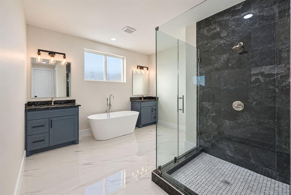 Dual charcoal-toned vanities with stone countertops, a freestanding soaking tub, and a glass-enclosed shower featuring dark stone wall tile and a mosaic-tile floor