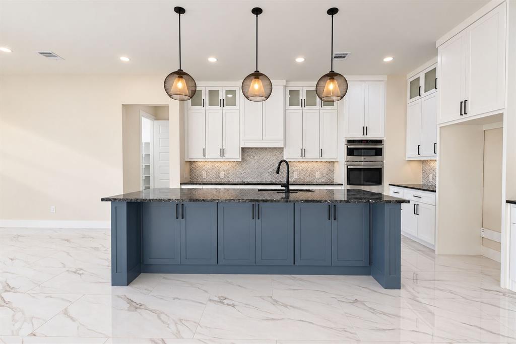 Kitchen featuring a central island with a dark stone countertop, white cabinetry with glass-front uppers, patterned tile backsplash, built-in stainless steel appliances, and triple pendant lighting