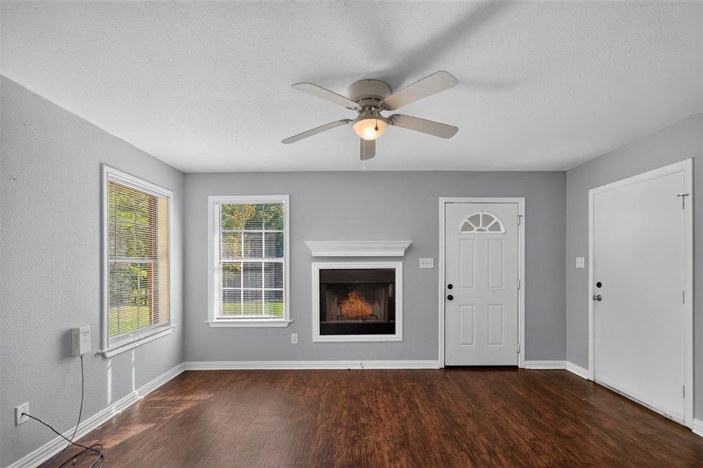 Living area featuring wood-finish flooring, a central fireplace with a decorative mantel, ceiling fan with integrated lighting, dual windows, and exterior door access