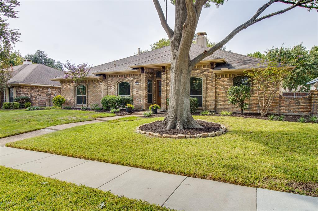 Brick exterior facade featuring an arched entryway, stone-bordered landscaping, prominent mature tree, and a concrete walkway