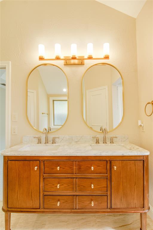 Double vanity featuring a light-toned stone countertop with dual undermount sinks, wood-finish cabinetry with brass hardware, dual oval mirrors, and a five-light gold-finish fixture