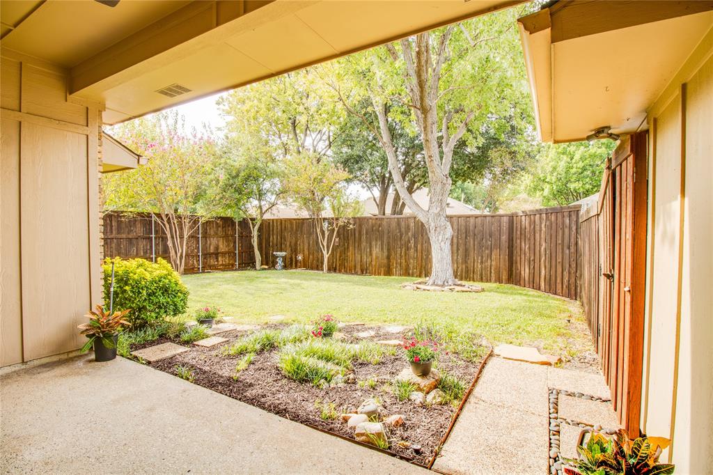Covered concrete patio with a landscaped garden border, enclosed backyard featuring a grass lawn, mature trees, and a full-height wood privacy fence