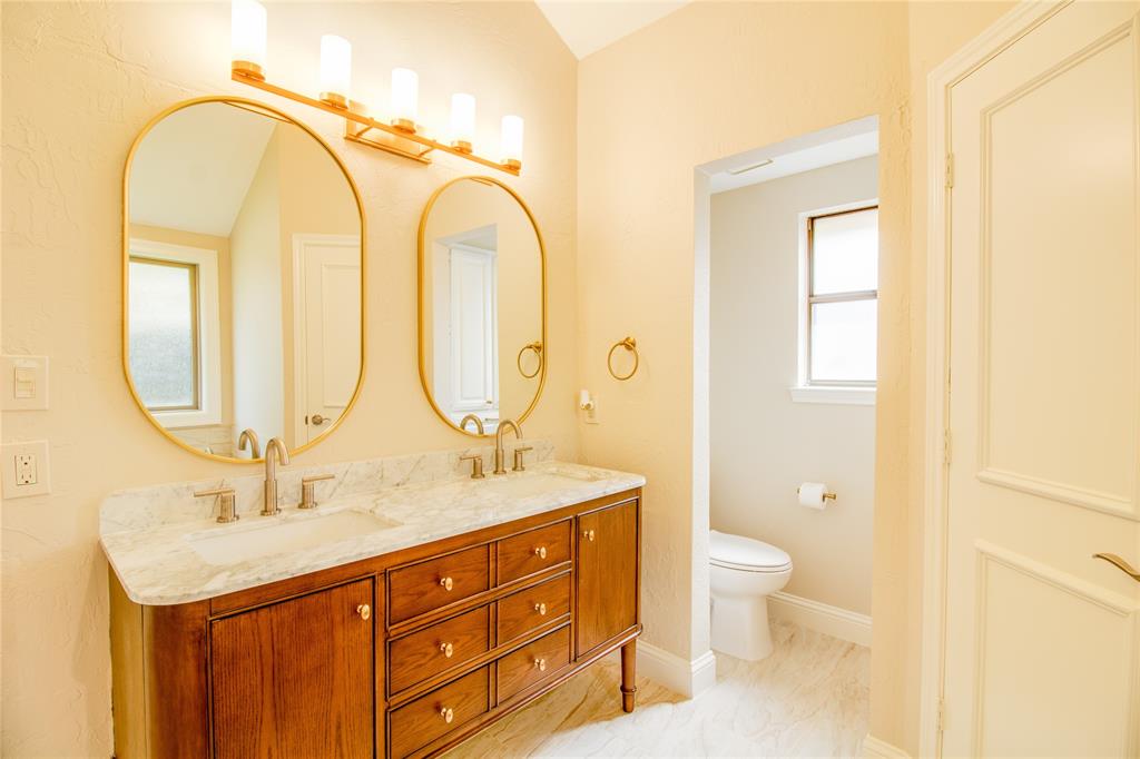 Double vanity with stone countertop, undermount sinks, wood cabinetry, oval wall-mounted mirrors, and four-light gold-toned fixture