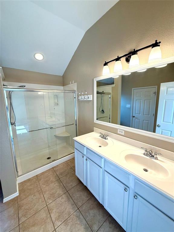 Bathroom featuring a double-sink vanity with white cabinetry, a glass-enclosed shower, a large wall-mounted mirror, and tile flooring under a vaulted ceiling