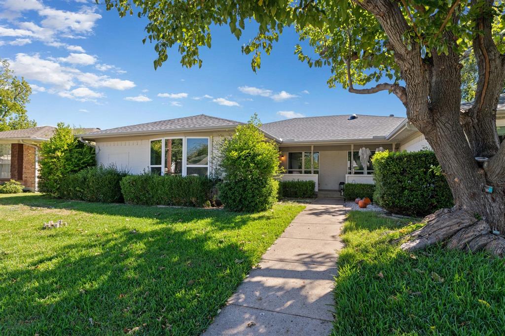 Single-story exterior featuring a light-toned stucco façade, a multi-pane bay window, a covered front entryway, and mature landscaping including a prominent front yard tree Single-story exterior featuring a light-toned stucco façade, a multi-pane bay window, a covered front entryway, and mature landscaping including a prominent front yard tree
