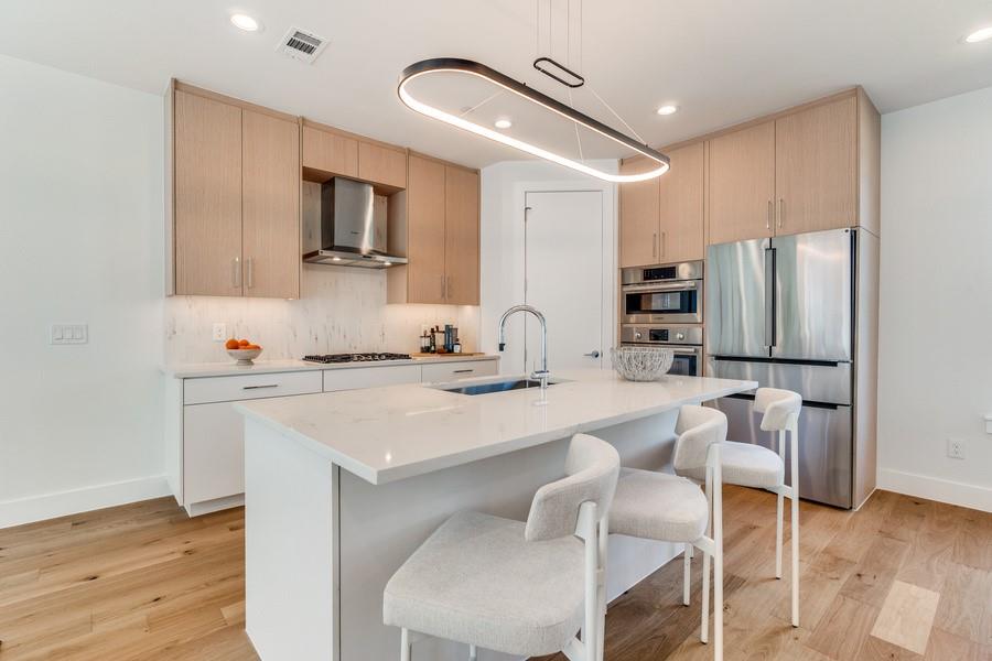 Kitchen featuring wood-finish cabinetry, stone-surface countertops, stainless steel appliances, a center island with an undermount sink, and minimalist pendant lighting