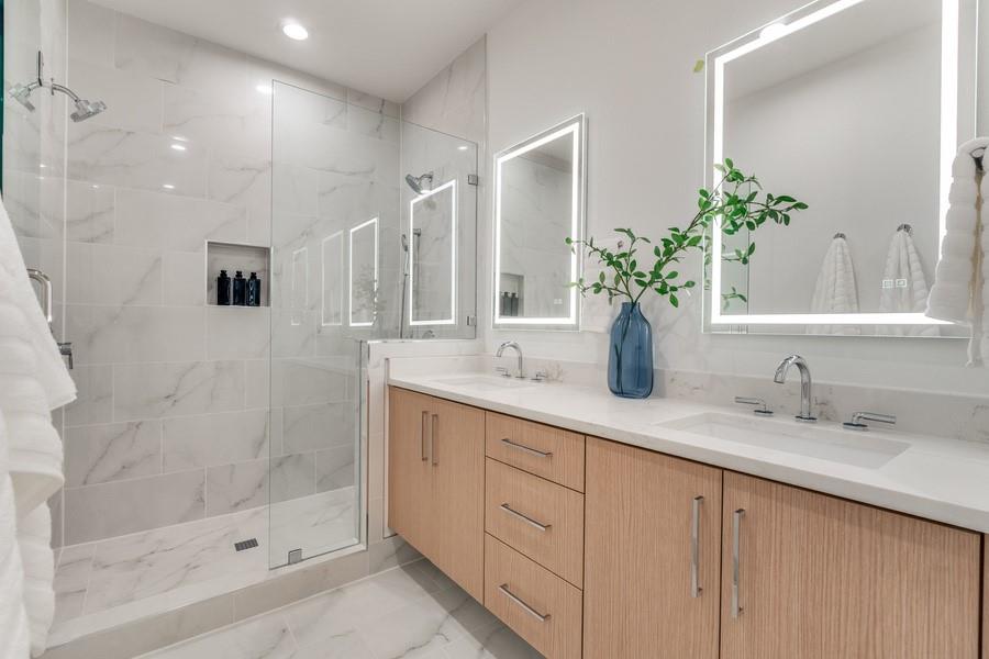 Dual-vanity cabinet with wood-finish cabinetry, solid-surface countertop, integrated rectangular sinks, frameless glass-enclosed shower featuring a recessed niche, and wall-mounted, backlit mirrors