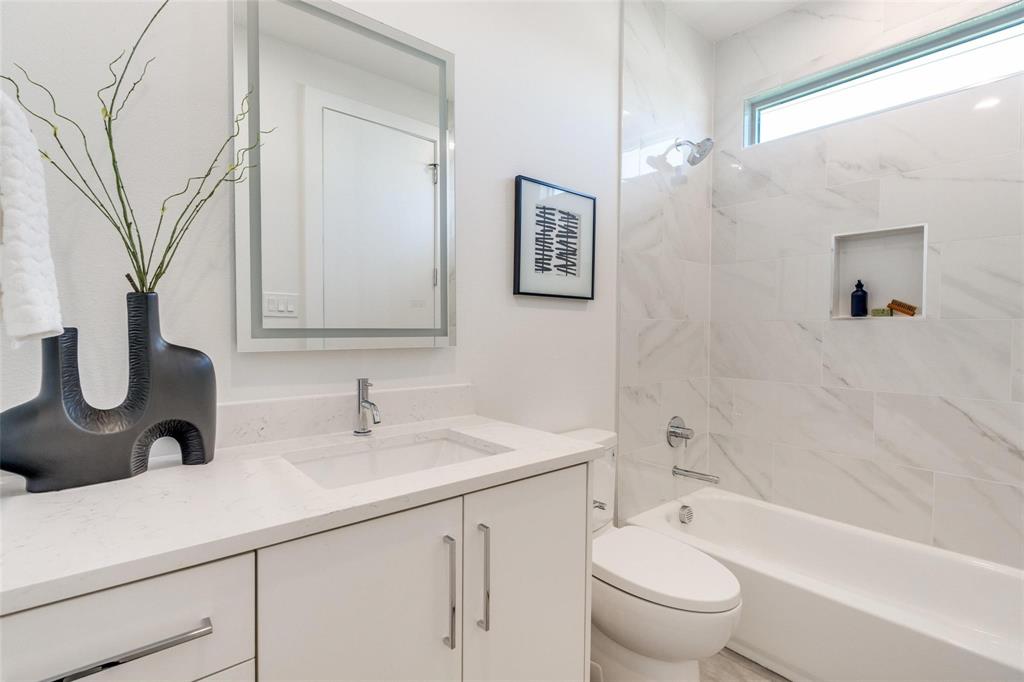 Bathroom featuring a white vanity with a light-toned countertop, integrated rectangular sink, wall-mounted backlit mirror, and a tub-shower combination with floor-to-ceiling stone-look tile, built-in shelving, and transom window