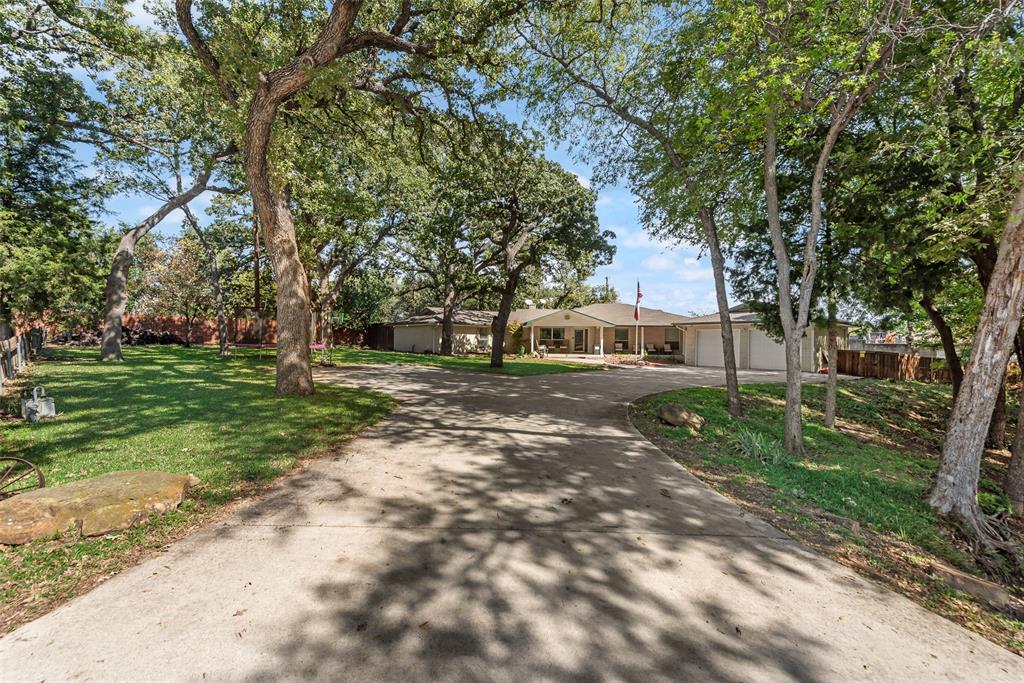 Single-story exterior featuring a concrete driveway, two-car garage, established mature trees, and natural stone landscaping elements Single-story exterior featuring a concrete driveway, two-car garage, established mature trees, and natural stone landscaping elements