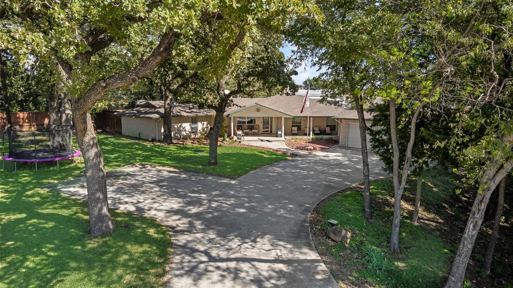 Single-story exterior featuring a brick facade, covered front porch, attached garage, curved concrete driveway, and established mature trees Single-story exterior featuring a brick facade, covered front porch, attached garage, curved concrete driveway, and established mature trees