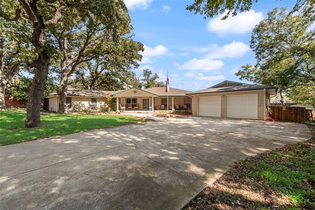 Single-story brick exterior featuring a covered front porch, an attached two-car garage, expansive concrete driveway, and mature trees on a landscaped lot Single-story brick exterior featuring a covered front porch, an attached two-car garage, expansive concrete driveway, and mature trees on a landscaped lot