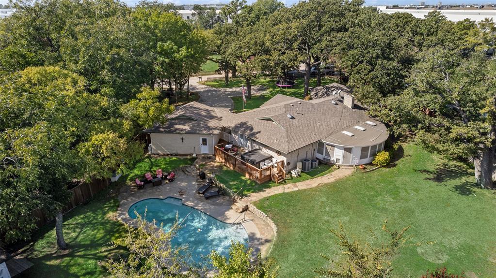Aerial perspective of a sprawling property featuring a swimming pool, stone-surfaced patio, elevated wood decking, and mature trees across a grass landscape