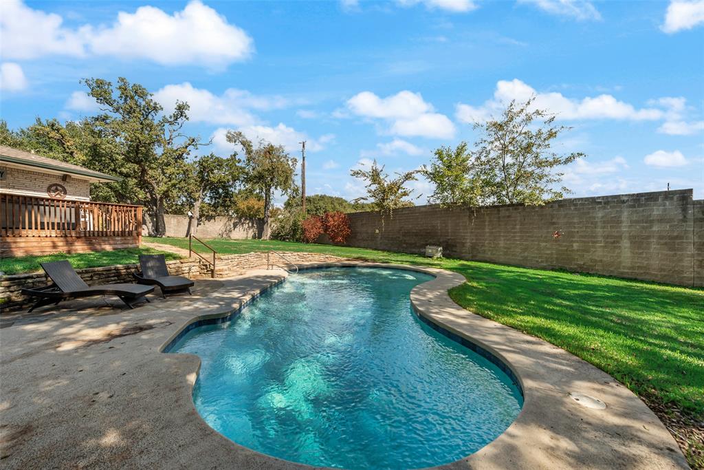 Free-form swimming pool with concrete decking, stone retaining wall, raised wooden deck, and expansive grass lawn enclosed by a masonry wall