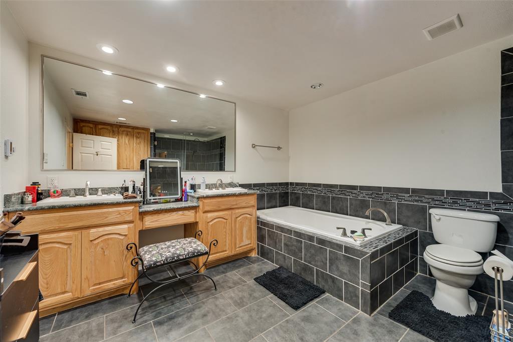 Dual-sink wood vanity with stone-finish countertops, deep soaking tub featuring dark tile surround, large wall-mounted mirror, and tile flooring