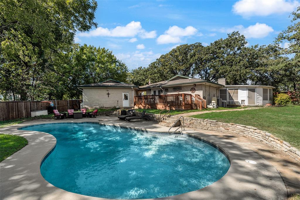 Inground swimming pool with concrete decking, stone retaining wall, raised wooden deck, and mature trees