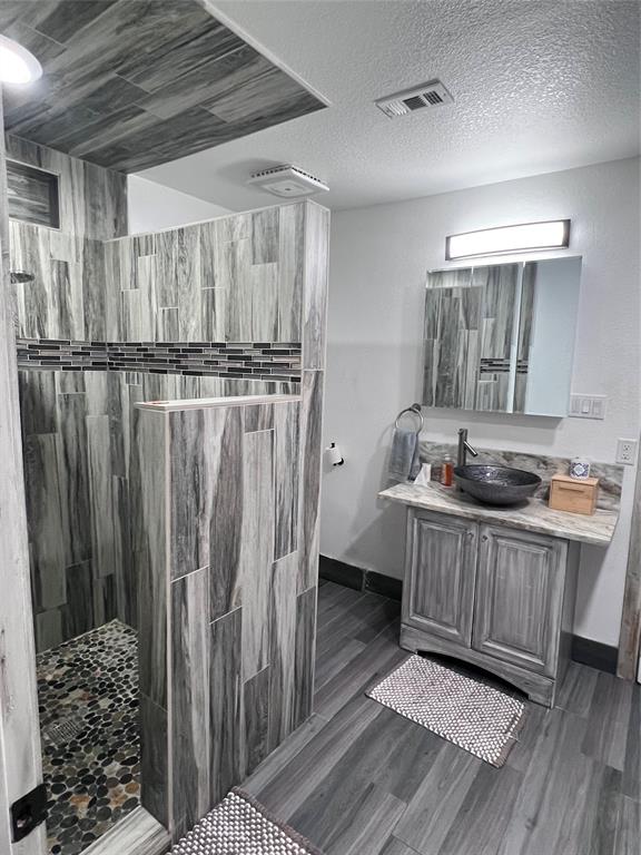 Walk-in shower with stone-tile flooring and vertical-pattern tile surround, vanity with decorative stone-finish countertop, vessel sink, and wall-mounted vanity lighting