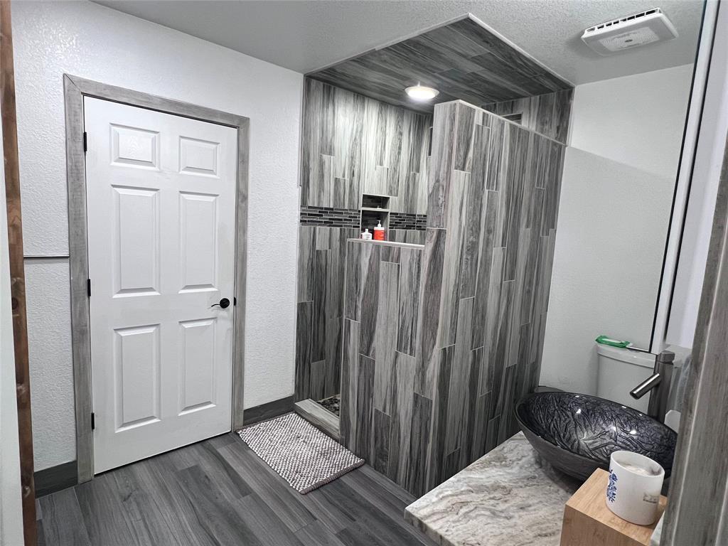 Walk-in shower with vertical wood-finish tile and recessed shelving, stone-finish countertop, vessel sink, and wood-finish flooring