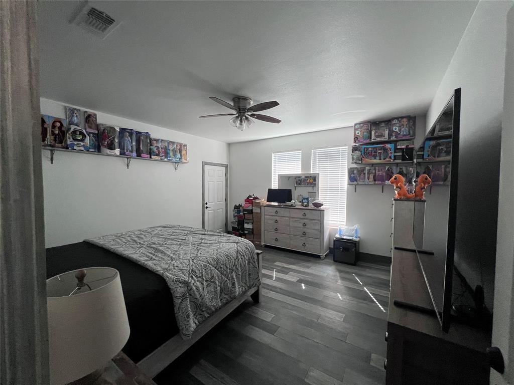 Room featuring wood-finish flooring, a ceiling fan with integrated lighting, dual windows with horizontal blinds, and two wall-mounted storage shelves