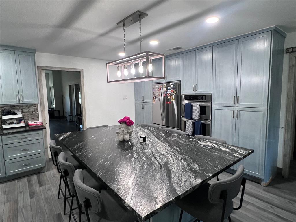 Kitchen featuring custom light blue cabinetry, a large stone-topped island, a linear multi-bulb pendant light fixture, stainless steel double wall ovens, and wood-finish flooring Kitchen featuring custom light blue cabinetry, a large stone-topped island, a linear multi-bulb pendant light fixture, stainless steel double wall ovens, and wood-finish flooring
