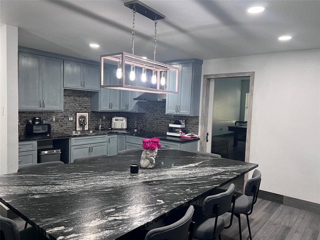 Kitchen featuring a large island with a stone countertop, cabinetry with light blue finish, rectangular pendant light fixture, stone-style backsplash, and wood-finish flooring Kitchen featuring a large island with a stone countertop, cabinetry with light blue finish, rectangular pendant light fixture, stone-style backsplash, and wood-finish flooring