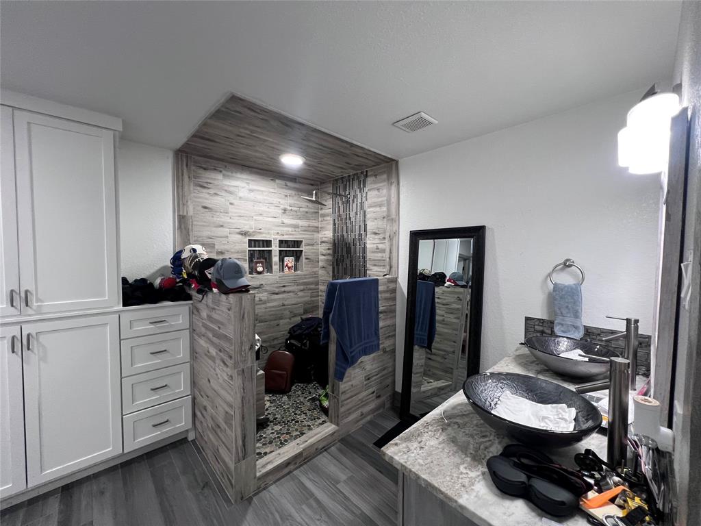 Double vanity featuring stone-patterned countertops, dual vessel sinks with metallic fixtures, full-height white cabinetry with drawer storage, and a walk-in shower with wood-finish wall tile and stone flooring