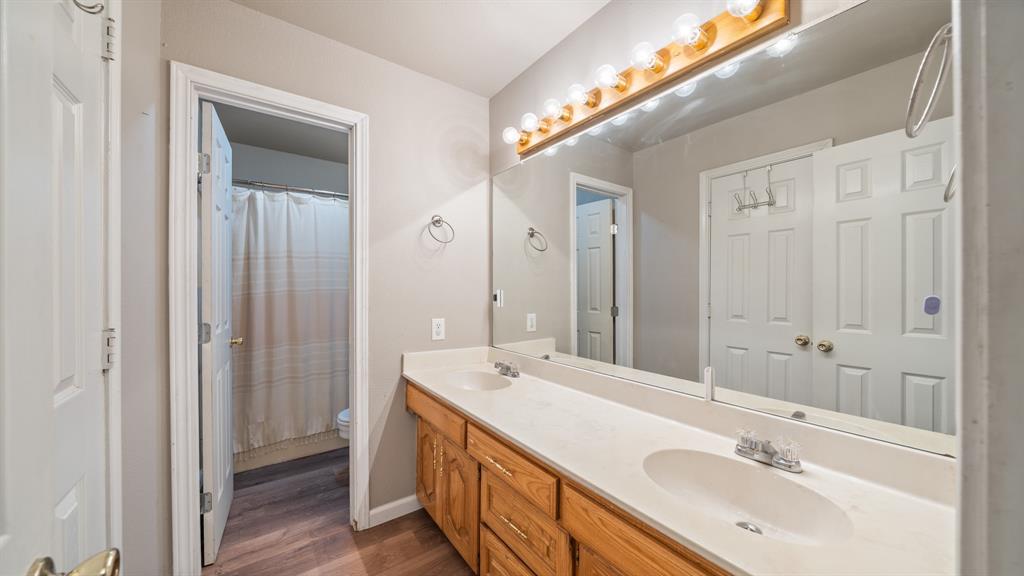 Bathroom vanity featuring a light-toned countertop with integrated dual sinks, wooden cabinetry, a full-width wall mirror, and a linear light fixture